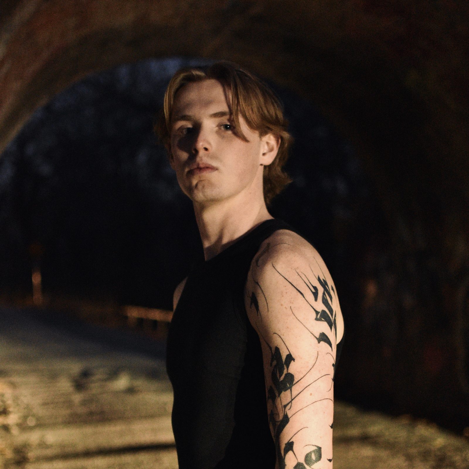Aidan April looking over his shoulder in a dark brick tunnel, with a black calligraphic tattoo across his arm